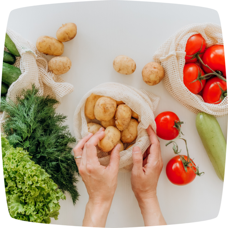 Auf einem weißen Untergrund sind verschiedene Gemüsesorten und Lebensmittel ausgebreitet. Eine Hand greift nach den Tomaten in der Mitte. Die Lebensmittel sind in wiederverwendbaren Netzbeuteln eingepackt.