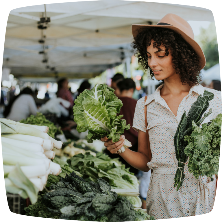 Eine junge Frau mit braunen Locken steht vor einem Gemüsestand auf dem Markt. Sie trägt ein weißes Kleid, einen braunen Hut und sucht sich Gemüse aus. Vor ihr auf dem Stand liegen Lauch und Grünkohl.