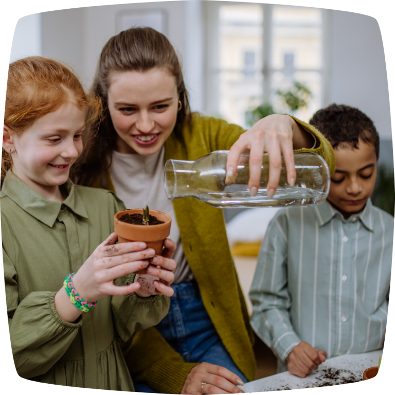 Eine Lehrerin und zwei Kinder gießen zusammen eine kleine Pflanze in einem Ton-Topf. Im Vordergrund befindet sich ein Mädchen, welches den Topf hält. Die Lehrerin gießt mit einer Glaskaraffe Wasser in den Topf.