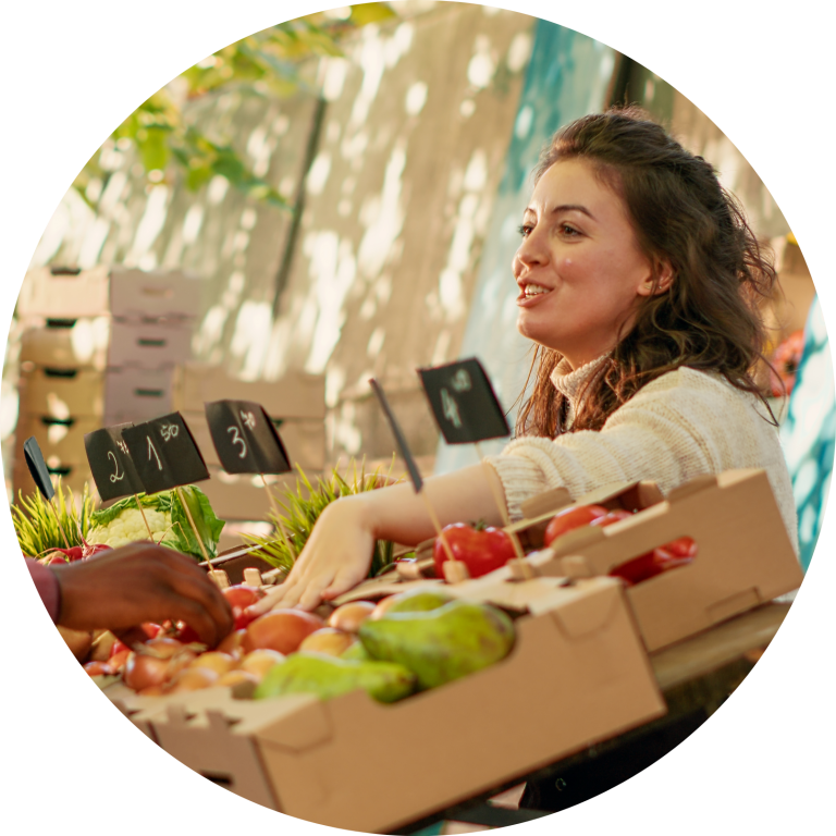 Eine junge Frau verkauft an ihrem Stand auf dem Markt Obst und Gemüse. Sie trägt einen weißen Rollkragenpullover und steht hinter den saisonalen Waren, welche in braunen Obstkisten aus Pappe präsentiert werden.
